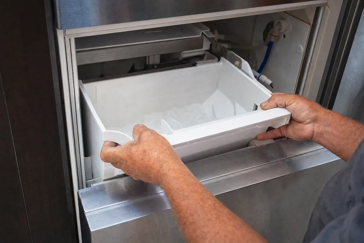 Two hands lifting the white ice bin out of a stainless undercounter ice machine, the mould and cutting grid visible above it