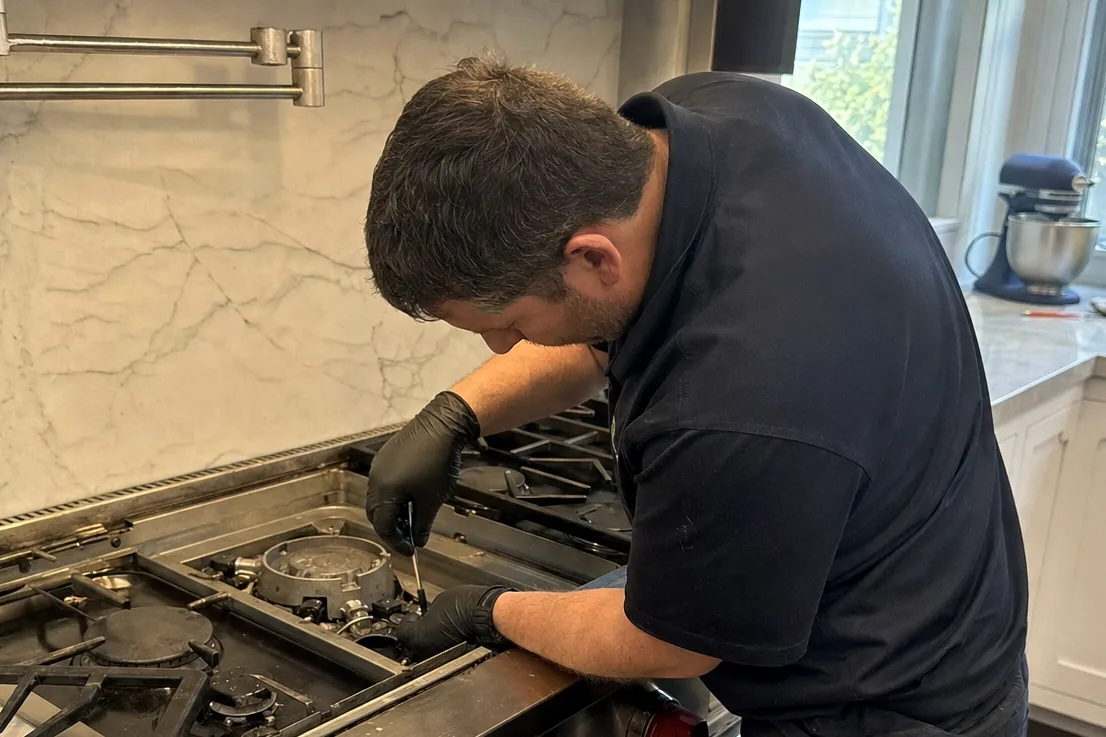 A technician bent over a stainless range in a working kitchen, one burner disassembled beside him