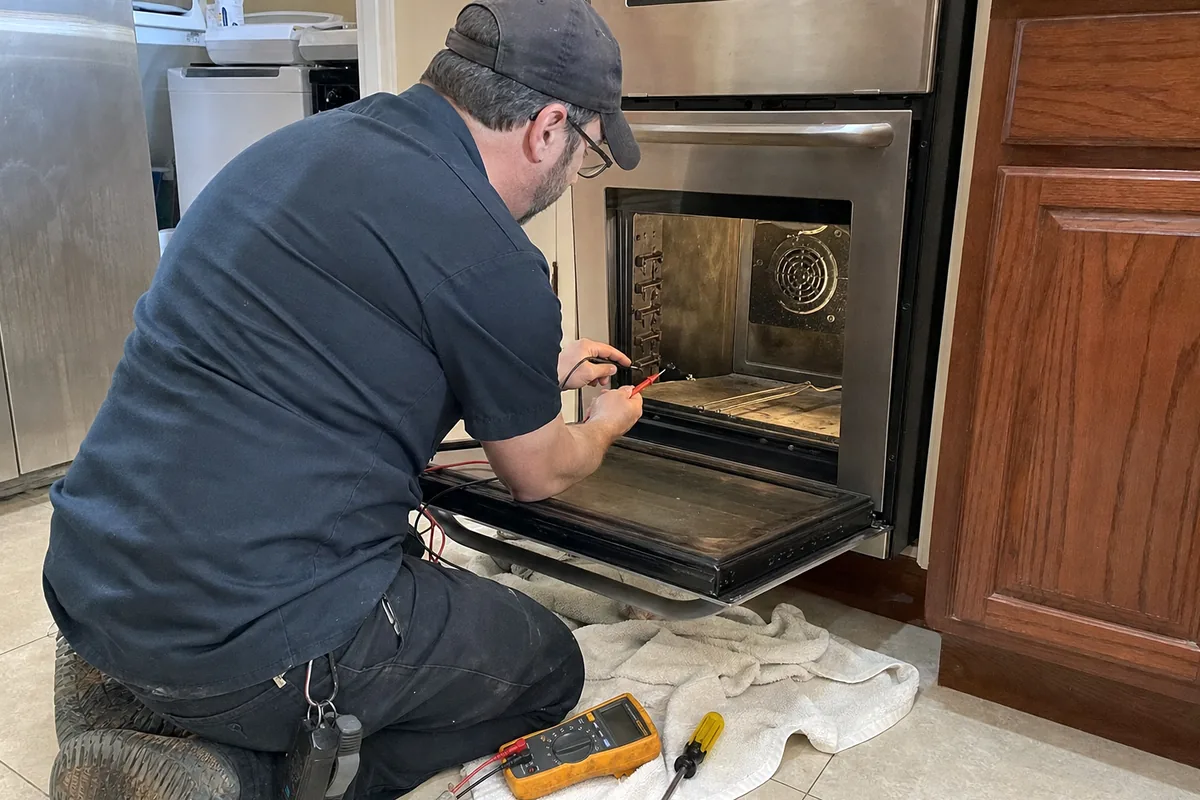 The open door of a stainless wall oven seen from the side, the technician reaching into the upper cavity