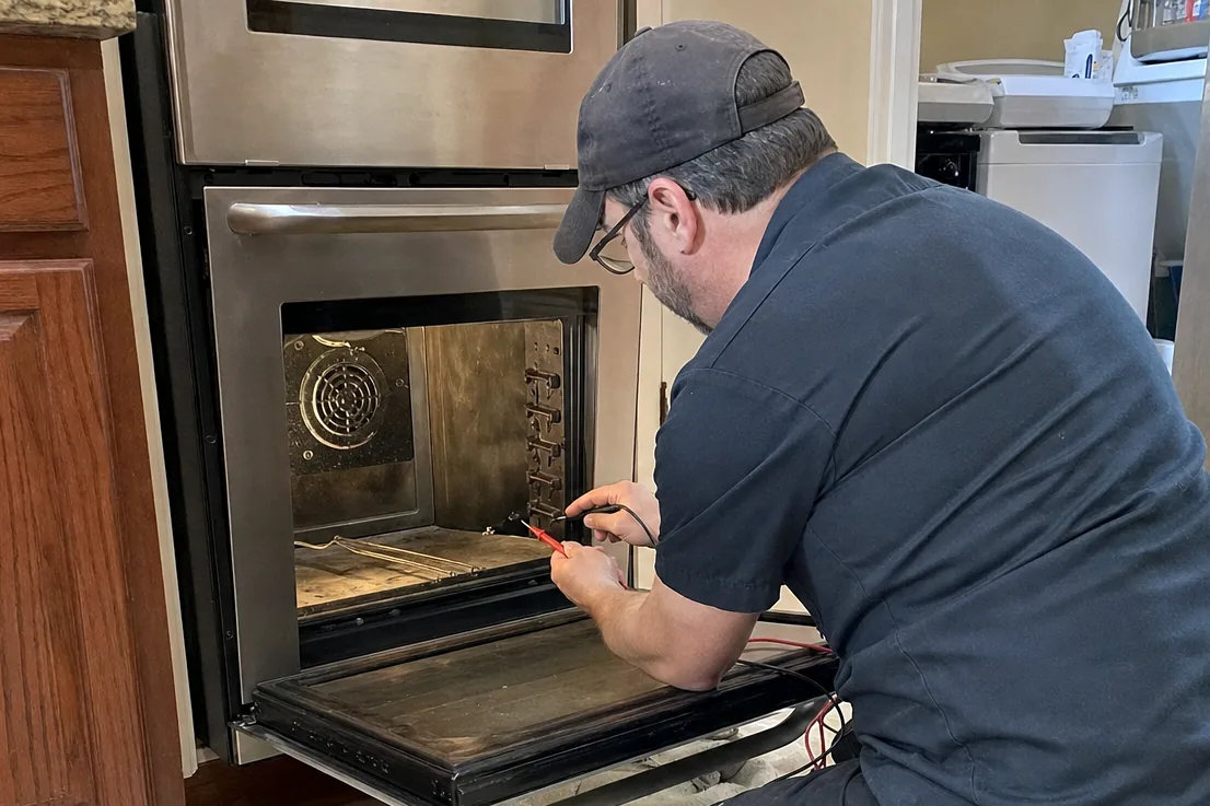 A technician working at the open door of a built-in oven, meter and drivers on the cloth beside his knee